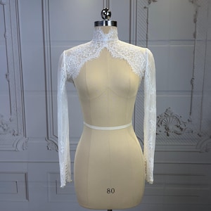 May include: A white lace bolero with long sleeves, a high neck, and scalloped edges. The sleeves feature small white buttons at the cuffs. The garment is displayed on a beige mannequin torso with the number 80 printed on the front.