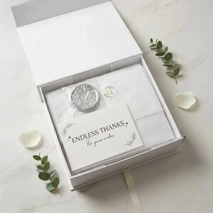 May include: A white gift box with an open lid, revealing a silver item in a plastic wrapper, a card with the text "ENDLESS THANKS For your order", and a small sticker. The box is lined with white tissue paper and decorated with greenery.