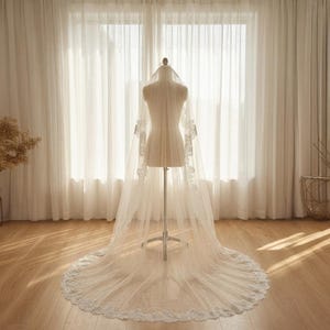 May include: A long, white bridal veil with delicate lace detailing along the edges and sleeves. The veil is displayed on a mannequin, showcasing its flowing design. The background features sheer white curtains and natural light.