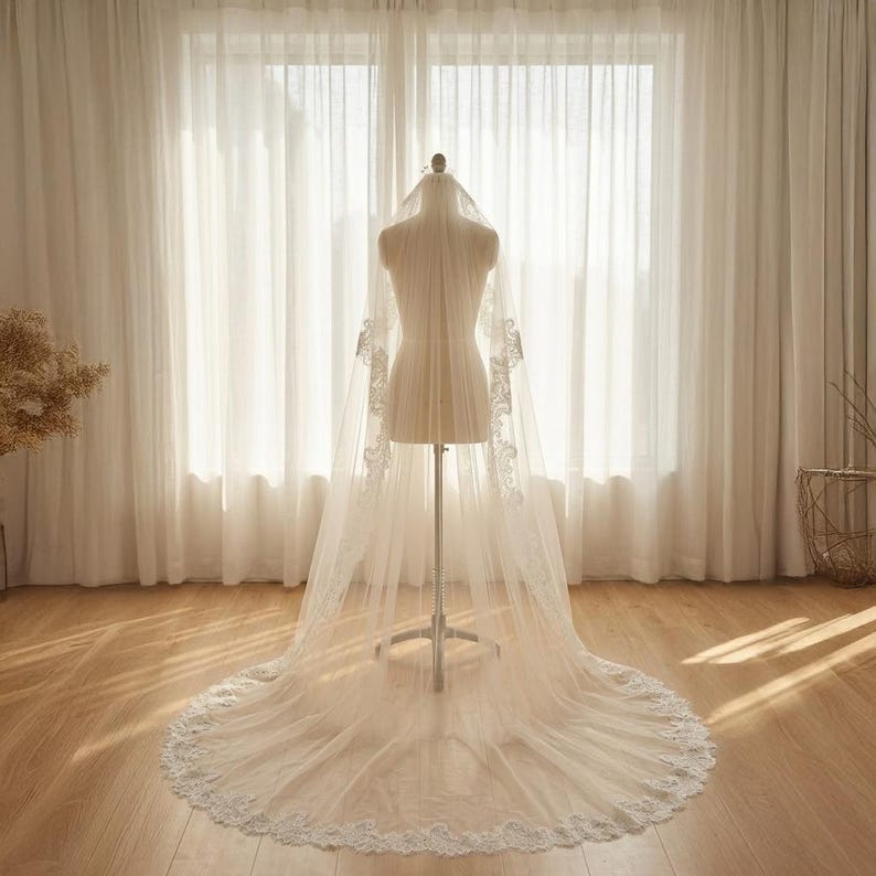 May include: A long, white bridal veil with delicate lace detailing along the edges and sleeves. The veil is displayed on a mannequin, showcasing its flowing design. The background features sheer white curtains and natural light.