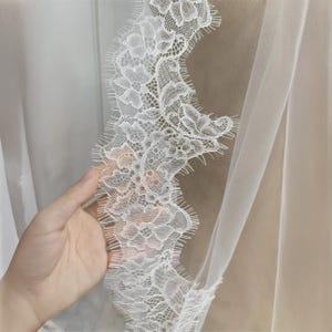 May include: Close-up of a white lace trim with a delicate floral pattern. The lace is edged with a scalloped design. The fabric is sheer and appears to be part of a veil or curtain.