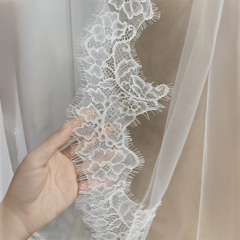May include: Close-up of a white lace trim with a delicate floral pattern. The lace is edged with a scalloped design. The fabric is sheer and appears to be part of a veil or curtain.