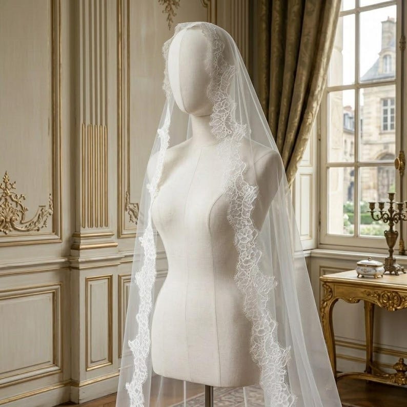 May include: A white bridal veil with delicate lace trim is displayed on a mannequin. The veil is sheer and drapes gracefully, showcasing the intricate floral lace along the edges. The mannequin is in a room with ornate wall paneling and a window.