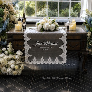 May include: A rectangular white lace table runner featuring intricate floral edges and the printed text Just Married, THOMAS & OLIVIA, est 2026. The runner measures 30 inches by 50 inches and is displayed on a dark wooden table with floral arrangements.
