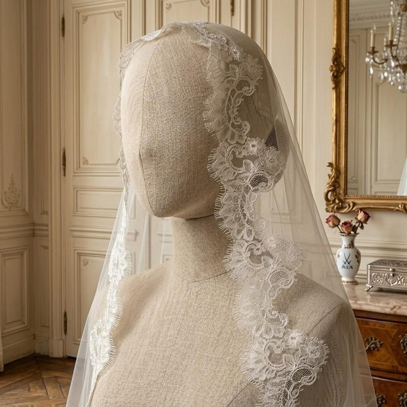 May include: A sheer white bridal veil with intricate lace detailing along the edges. The veil is draped over a mannequin head, showcasing the delicate floral patterns and the veil's flowing design. The veil is a classic wedding accessory.
