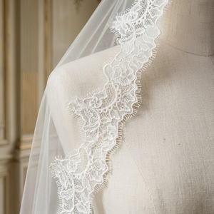 May include: A white bridal veil with a delicate floral lace trim. The lace is intricately designed with detailed patterns and a scalloped edge. The veil is draped over a mannequin, showcasing the soft, flowing fabric and the intricate lace detail.