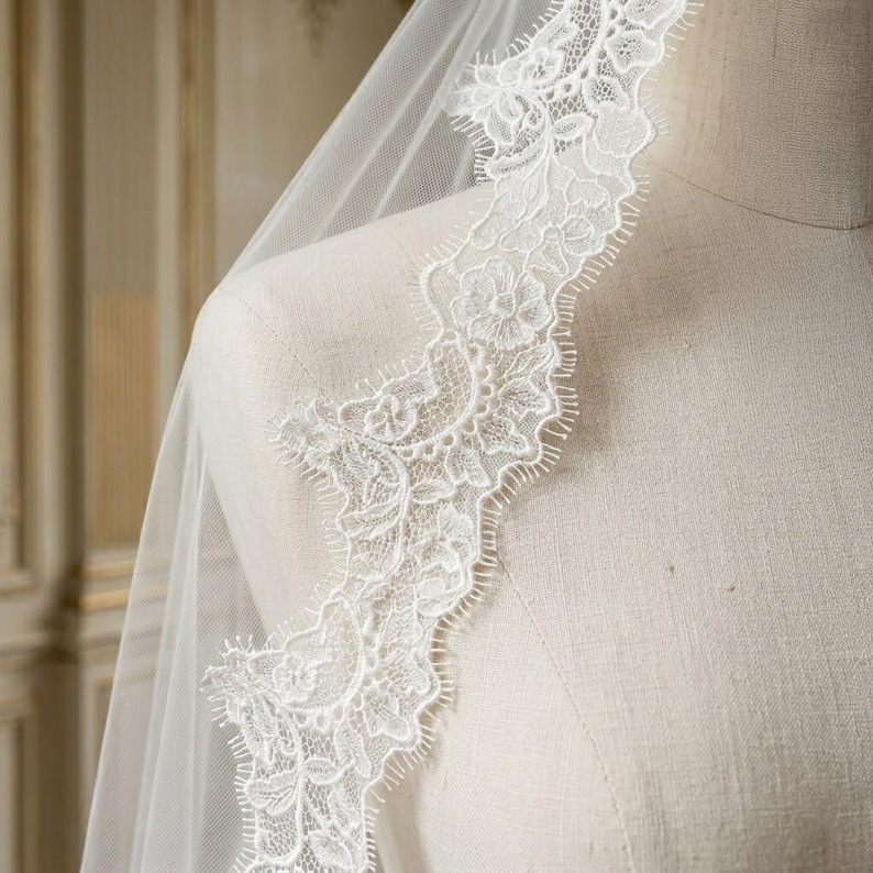 May include: A white bridal veil with a delicate floral lace trim. The lace is intricately designed with detailed patterns and a scalloped edge. The veil is draped over a mannequin, showcasing the soft, flowing fabric and the intricate lace detail.