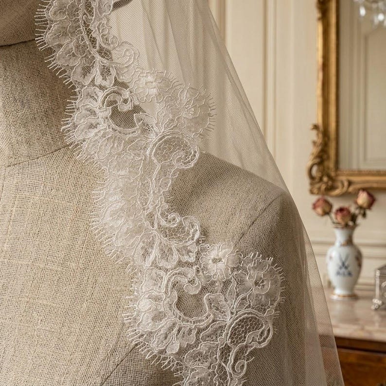 May include: A close-up of a bridal veil with intricate white lace detailing. The lace trim features floral and scroll patterns along the edge of the sheer fabric. The veil is draped over a mannequin, showcasing the delicate design.