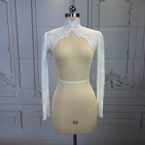 May include: A white lace bolero with long sleeves and a high neck, featuring intricate floral patterns and scalloped edges. The sleeves are finished with a row of small white pearl buttons at the cuffs. The garment is displayed on a beige mannequin.