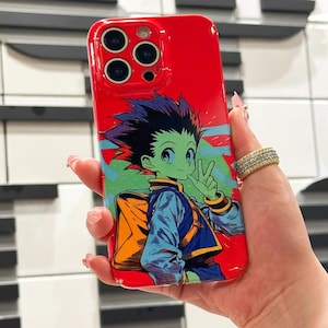 以下が含まれることがあります： 赤い携帯電話ケース。アニメ風のイラストで、黒いとげのある髪、緑色の目、青い衣装のキャラクターが描かれています。キャラクターはピースサインをしています。ケースは光沢仕上げで、カメラの切り抜きがあります。