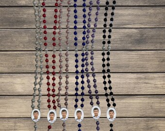 May include: Six different colored rosaries with a silver cross and a photo charm. The rosaries are white, red, pink, blue, purple, and black.