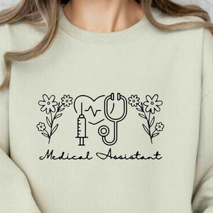 Puede incluir: Una sudadera de color beige claro con un diseño gráfico negro de un corazón con un estetoscopio y una jeringa, rodeado de flores. El texto "Medical Assistant" está impreso debajo del gráfico.