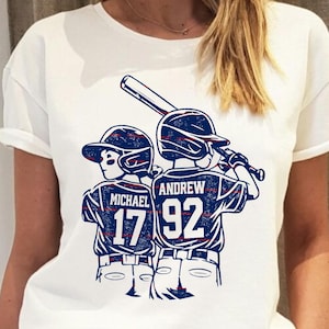 Op de afbeelding: Wit t-shirt met een honkbalafbeelding. Het ontwerp toont twee honkbalspelers in blauw-witte uniformen, met de namen "MICHAEL" en "ANDREW" en de nummers "17" en "92" op de shirts. Een honkbalknuppel is ook zichtbaar.