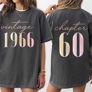 May include: Dark gray t-shirt with the text "vintage 1966" on the front and "chapter 60" on the back. The numbers are in a combination of beige and pink. The shirt has a relaxed fit.