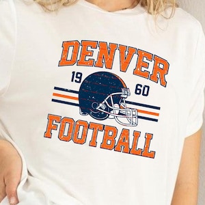 May include: White t-shirt with a Denver Football graphic. The design features the word "DENVER" in orange block letters above a navy blue football helmet, the numbers "19" and "60", and the word "FOOTBALL" in orange.