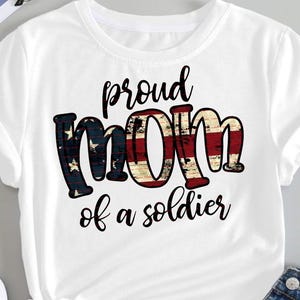 Puede incluir: Camiseta blanca con la frase "proud MOM of a soldier" en letras negras. La palabra "MOM" está diseñada con un patrón de bandera americana desgastada. La camiseta se combina con vaqueros azules y zapatillas blancas.