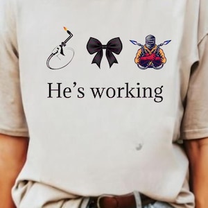 May include: A white t-shirt with a graphic of a welding torch, a black bow, and a welder with a welding torch. The text "He's working" is printed below the graphic.