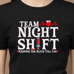 May include: Black t-shirt featuring the text "TEAM NIGHT SHIFT" in white, a red heart with a nurse's hat, and a syringe graphic. The phrase "KEEPING 'EM ALIVE TILL 7:05" is printed below the main text.