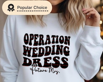 Puede incluir: Sudadera blanca con el texto "OPERATION WEDDING DRESS Future Mrs." en letras negras y rojas. La sudadera tiene cuello redondo y mangas largas. Una insignia de "Popular Choice" está en la esquina superior izquierda.