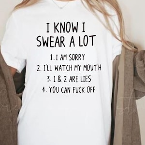 以下が含まれることがあります： 「I KNOW I SWEAR A LOT」の黒い文字と一連の文言がプリントされた白いTシャツ。「I AM SORRY」、「I'LL WATCH MY MOUTH」、「1 & 2 ARE LIES」、「YOU CAN FUCK OFF」のフレーズが含まれています。
