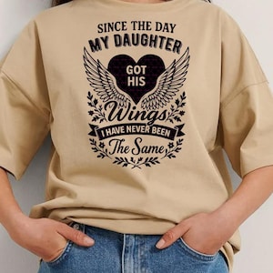 Puede incluir: Camiseta beige con texto negro: "SINCE THE DAY MY DAUGHTER GOT HIS WINGS I HAVE NEVER BEEN The Same". El diseño incluye un corazón y alas.