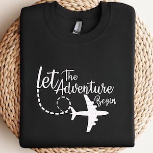 Puede incluir: Sudadera negra con el texto blanco "Let The Adventure Begin" y un gráfico de avión estilizado. El diseño incluye una línea de puntos, que sugiere una ruta de vuelo. La sudadera está doblada sobre una superficie circular tejida.