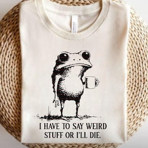 Peut inclure: T-shirt crème avec une illustration noire d'une grenouille tenant une tasse. La grenouille a de grands yeux et une expression légèrement grincheuse. Le texte "I HAVE TO SAY WEIRD STUFF OR I'LL DIE" est imprimé en dessous.