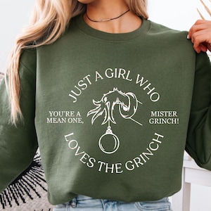 Könnte beinhalten: Olivgrünes Sweatshirt mit weißem Grinch-Motiv und Text. Der Text lautet: "Just a girl who loves the Grinch", ergänzt durch "You're a mean one" und "Mister Grinch!" in einem Kreis.