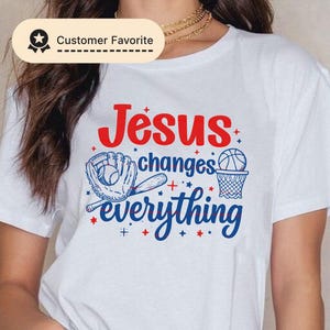 Könnte beinhalten: Weißes T-Shirt mit dem Text "Jesus changes everything" in Rot und Blau. Das Design umfasst einen Baseballhandschuh, einen Schläger, einen Basketball und einen Korb. Das T-Shirt hat einen Rundhalsausschnitt und kurze Ärmel.