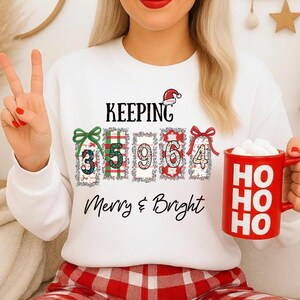 May include: White sweatshirt with the text "KEEPING Merry & Bright" and the numbers 3, 5, 9, 6, and 4 in decorative boxes. A red mug with "HO HO HO" is held in the hand. The person is wearing red and white plaid pants.