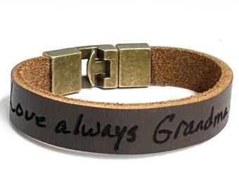 May include: A brown leather bracelet with the engraved words "Love always Grandma." The bracelet has a rectangular, antique brass-colored clasp. The leather band is approximately 0.75 inches wide.