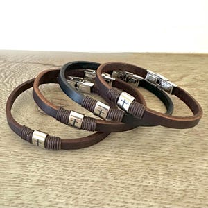May include: Four leather bracelets in shades of brown and black. Each bracelet features a silver-colored metal accent with a cross design. The bracelets have a simple, classic design with a clasp closure.