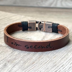 May include: Brown leather bracelet with the engraved text "I am second." The bracelet features a metal clasp and black cord accents. The bracelet is approximately 2 cm wide.