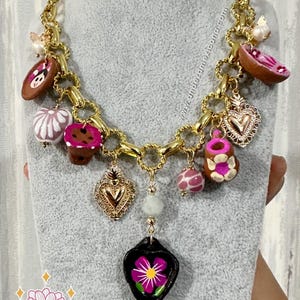 May include: A gold-toned chain necklace with various charms. Charms include a heart, a flower, and other decorative elements in pink, brown, and white. The necklace is displayed on a gray mannequin.