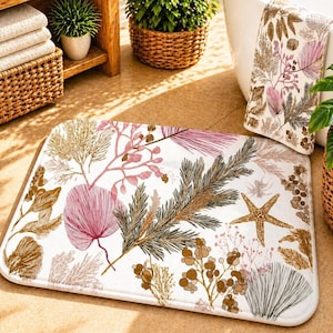 May include: White bath mat with a botanical design featuring pink, brown, and gold-colored illustrations of plants and a starfish. A matching towel is visible. The bath mat is rectangular and has rounded corners.
