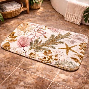 May include: Rectangular bath mat with a cream background and a botanical design. The design features illustrations of seaweed, starfish, and other sea life in shades of brown, pink, and green. The mat is placed on a tiled floor.