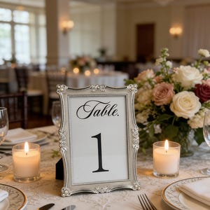 Könnte beinhalten: Tischdekoration für eine Hochzeit mit der Tischnummer 1 in einem silbernen Rahmen. Der Rahmen hat ein verziertes Design und das Wort "Table" steht über der Nummer. Zwei brennende Kerzen und ein Blumenarrangement befinden sich ebenfalls auf dem Tisch.