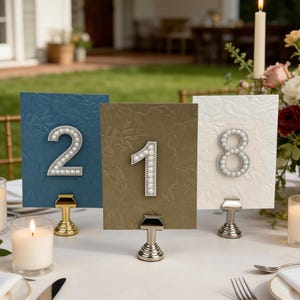 May include: Three table number cards with embossed floral patterns. Numbers 2, 1, and 8 are adorned with faux pearls or crystals. The cards are in blue, olive green, and white, each held by a metal stand. A candle and floral arrangement are in the background.