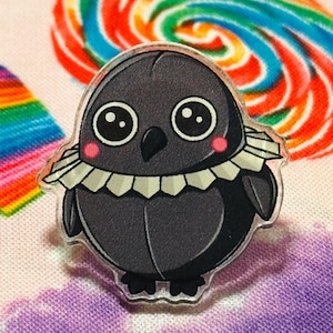May include: A black and white cartoon bird with a ruffled collar, big eyes, and pink cheeks. The bird is a pin made of clear acrylic.