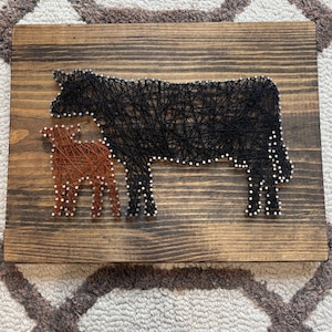 May include: A rustic wooden sign featuring a black cow and a smaller brown calf, both crafted with string art. The animals are outlined with white nail heads, creating a charming farmhouse aesthetic. The wood has a warm, natural finish.