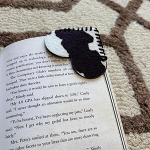 May include: A black and white cowhide pattern bookmark. The bookmark is made of felt and has a stitched border. It is resting on the pages of a book.