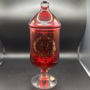 May include: A red glass jar with a gold inscription that reads "40th Anniversary 1975-2015". The jar has a lid and a pedestal base.