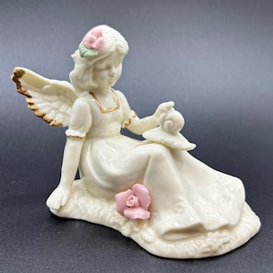 May include: A porcelain figurine of an angel with wings, wearing a white dress and a pink flower crown, sitting with a snail on a bed of flowers.