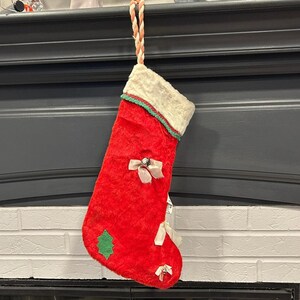 May include: A red Christmas stocking with a white cuff trimmed with green. The stocking features white bows with silver accents and a green felt Christmas tree. A twisted rope loop is attached for hanging.