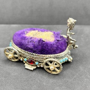 May include: A vintage silver-toned carriage-shaped pin cushion with a purple velvet cushion. The carriage has decorative wheels, turquoise accents, and a red jewel. A small figure sits at the back.