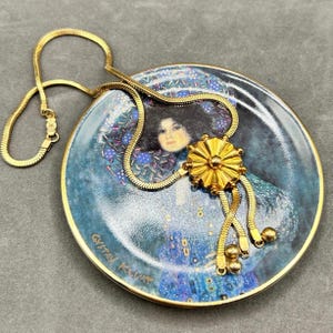 Peut inclure: Un collier doré avec un pendentif décoratif repose sur une assiette décorative. L'assiette présente un portrait avec des accents bleus et dorés. Le collier a un design de chaîne serpent et un pendentif en forme d'éclat de soleil avec des accents pendants.