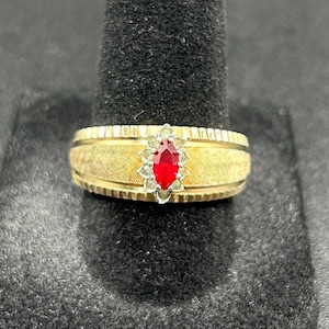 Puede incluir: Un anillo dorado con una banda texturizada y una gema roja central. La gema está rodeada de piedras más pequeñas y transparentes. El anillo se muestra sobre una superficie negra.