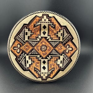 May include: A round, decorative plate with a geometric design in black, white, and orange. The plate has a raised edge and a smooth surface.