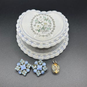 Könnte beinhalten: Eine herzförmige, weiße Keramik-Schmuckschatulle mit floralem Design, neben einem Paar hellblauer Strass-Ohrringe und einem kleinen Anhänger. Die Ohrringe und der Anhänger haben eine Vintage-Ästhetik. Die Box hat einen gewellten Rand.