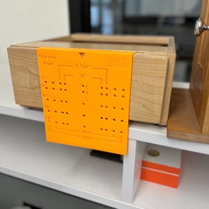 Puede incluir: Una plantilla naranja de plástico para tirador de cajón está sujeta a un cajón de madera. La plantilla tiene medidas en pulgadas y el texto "Drawer Pull" y "DRAFT SPEED". El cajón está sobre una estantería blanca.